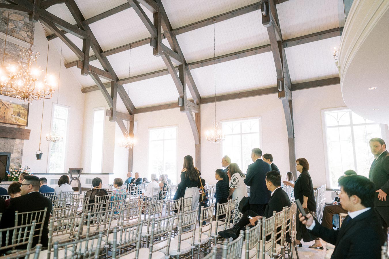 Wedding guests take their seats inside a bright chapel with timber beams and chandeliers at Ancaster Mill.