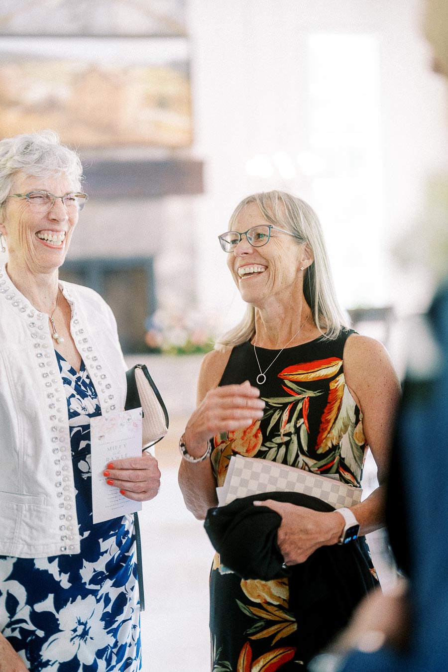 Two smiling women in glasses chat indoors at a wedding, one holding a Miffy and Rawle program.
