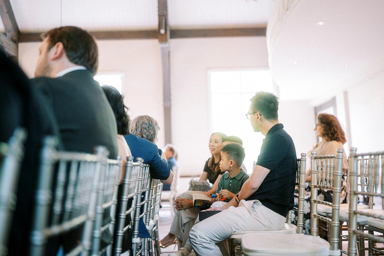 Guests seated on silver chairs inside a bright wedding ceremony room, including a family with a young child.