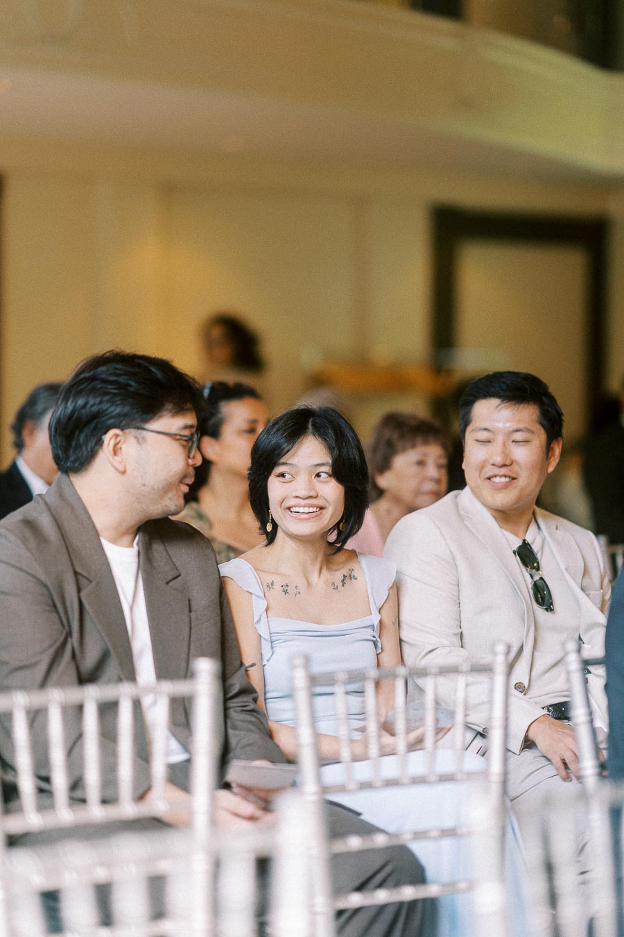 Guests seated on silver chairs chat and smile during an indoor wedding gathering.