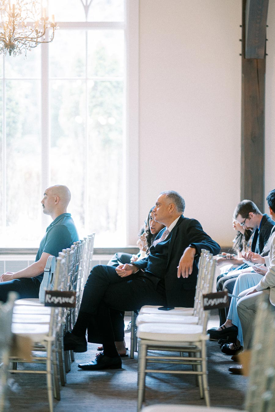 Wedding guests sit in clear chairs beside tall windows before the ceremony at Ancaster Mill.