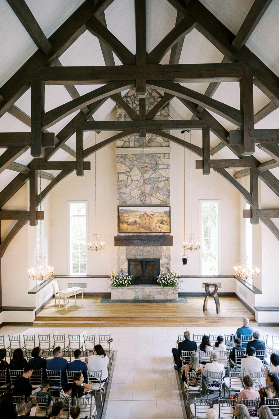 Guests seated in Ancaster Mill chapel beneath timber beams, chandeliers, and a stone fireplace before the ceremony