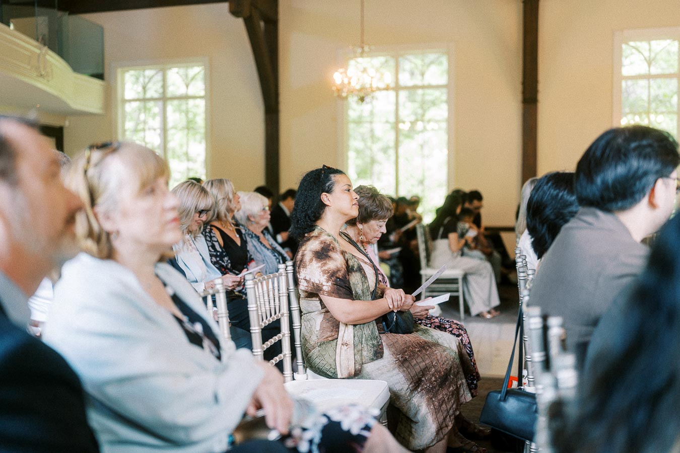 Wedding guests seated on silver chairs inside a bright chapel at Ancaster Mill, holding ceremony programs.