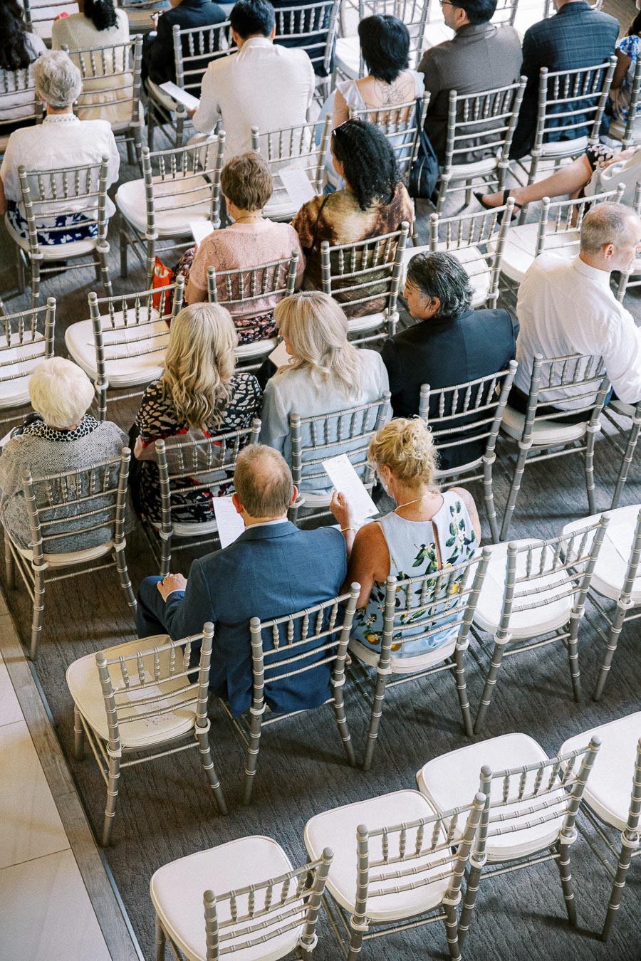 Wedding guests seated in silver chairs with ceremony programs before an indoor chapel ceremony.