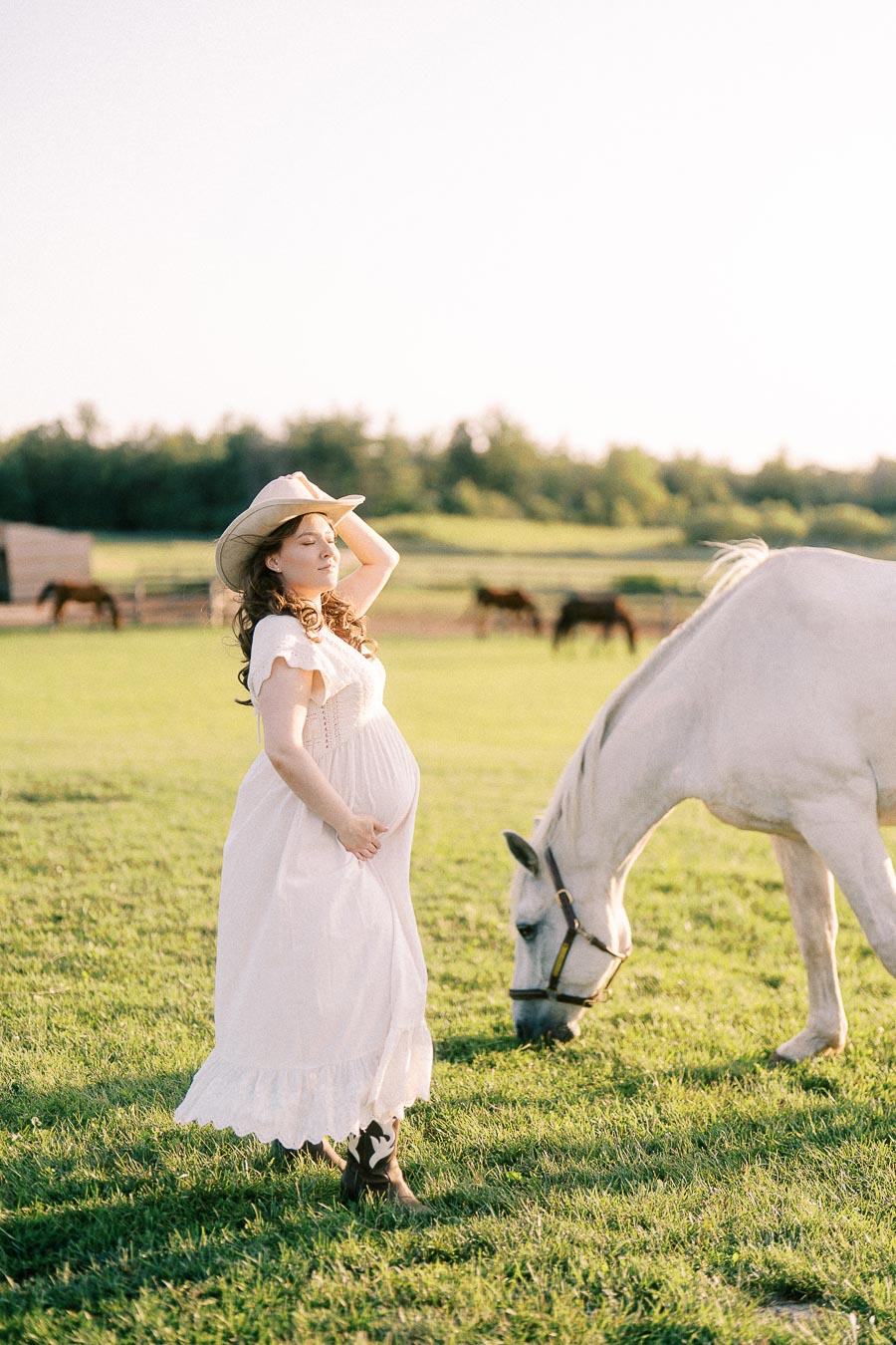 Pregnant woman in white dress and cowboy hat beside a grazing white horse