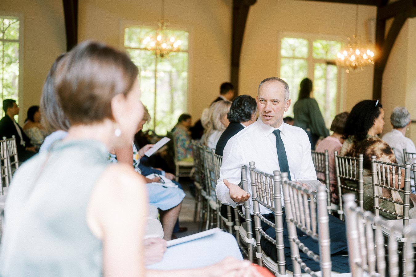 Guests seated in silver chairs chat inside a bright Ancaster Mill ceremony room with chandeliers and tall windows.