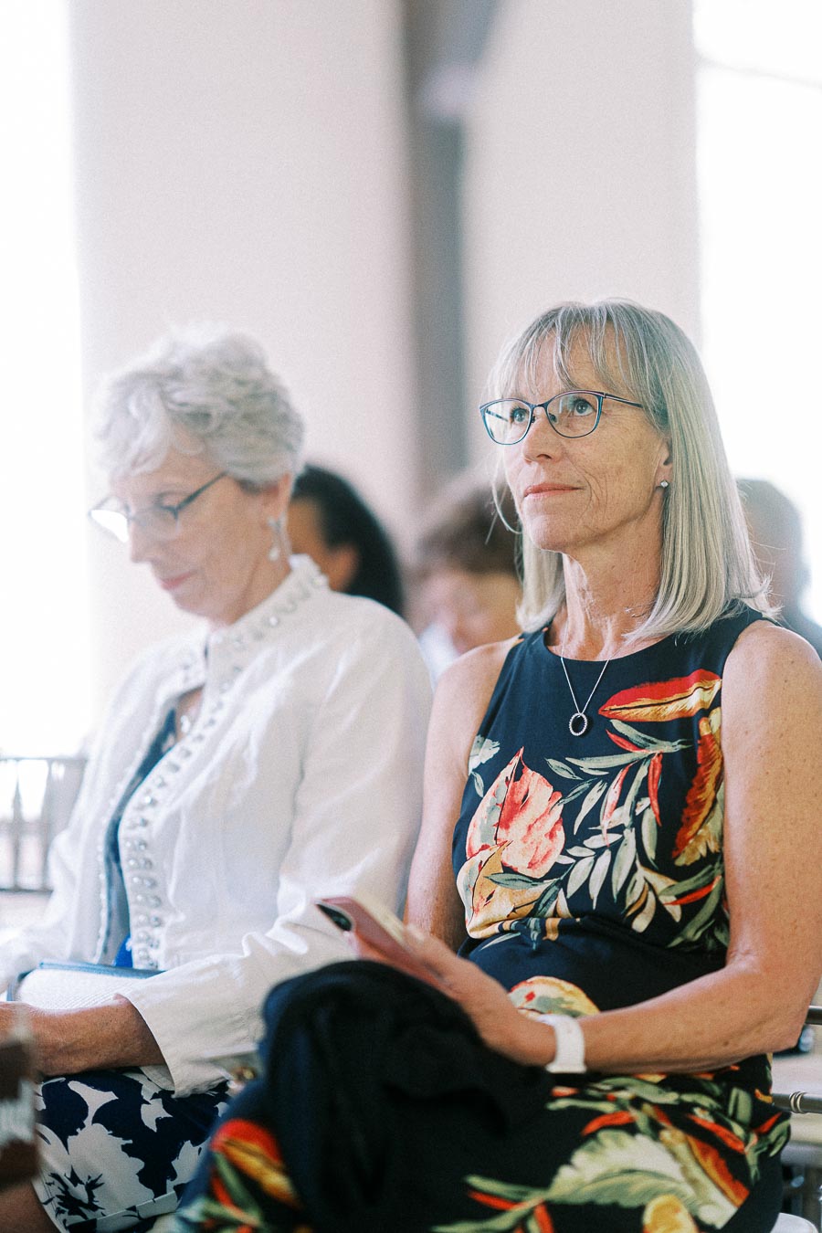 Two women seated indoors at a wedding ceremony, one in a floral dress looking ahead and another reading beside her.
