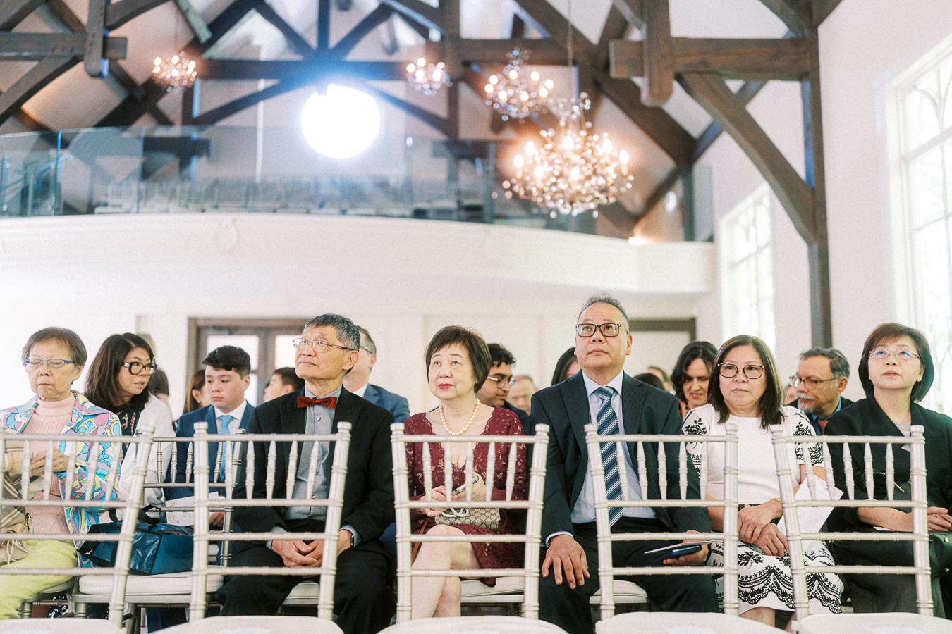 Wedding guests seated in white chairs beneath chandeliers and timber beams during an indoor ceremony