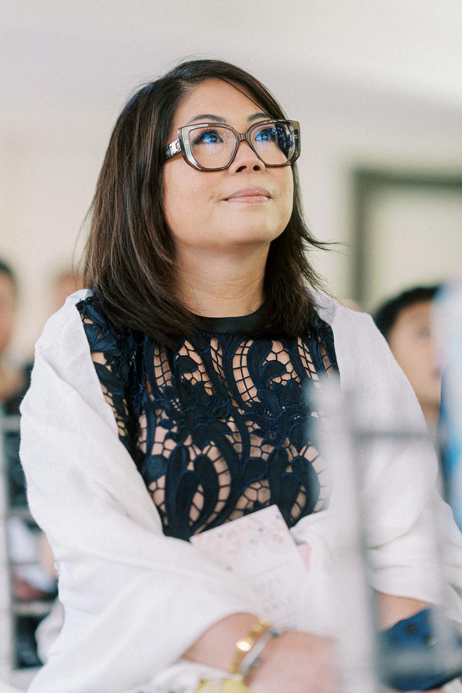 Woman in glasses and a white shawl listens attentively while seated at a wedding reception
