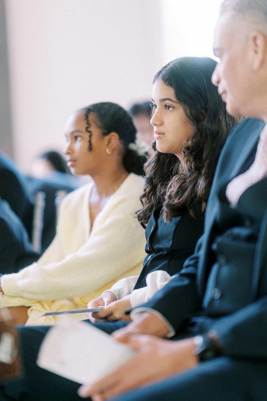 Guests seated in a bright ceremony space, holding programs and watching attentively from the pews.
