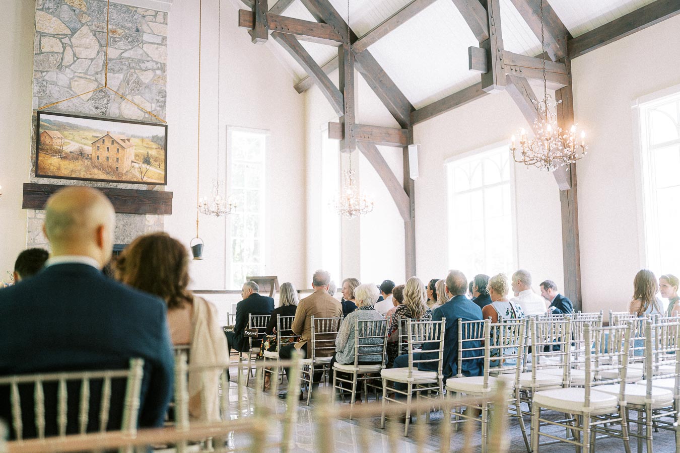 Wedding guests seated in a bright chapel with timber beams, chandeliers, stone fireplace, and silver chairs.
