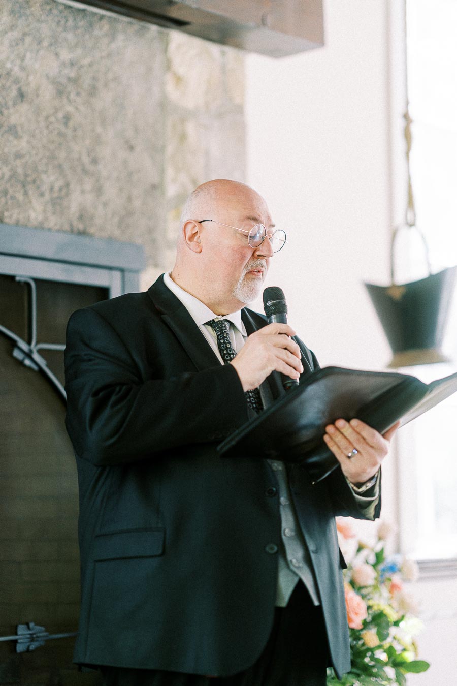 Wedding officiant in a black suit reads from a folder into a microphone during an indoor ceremony.