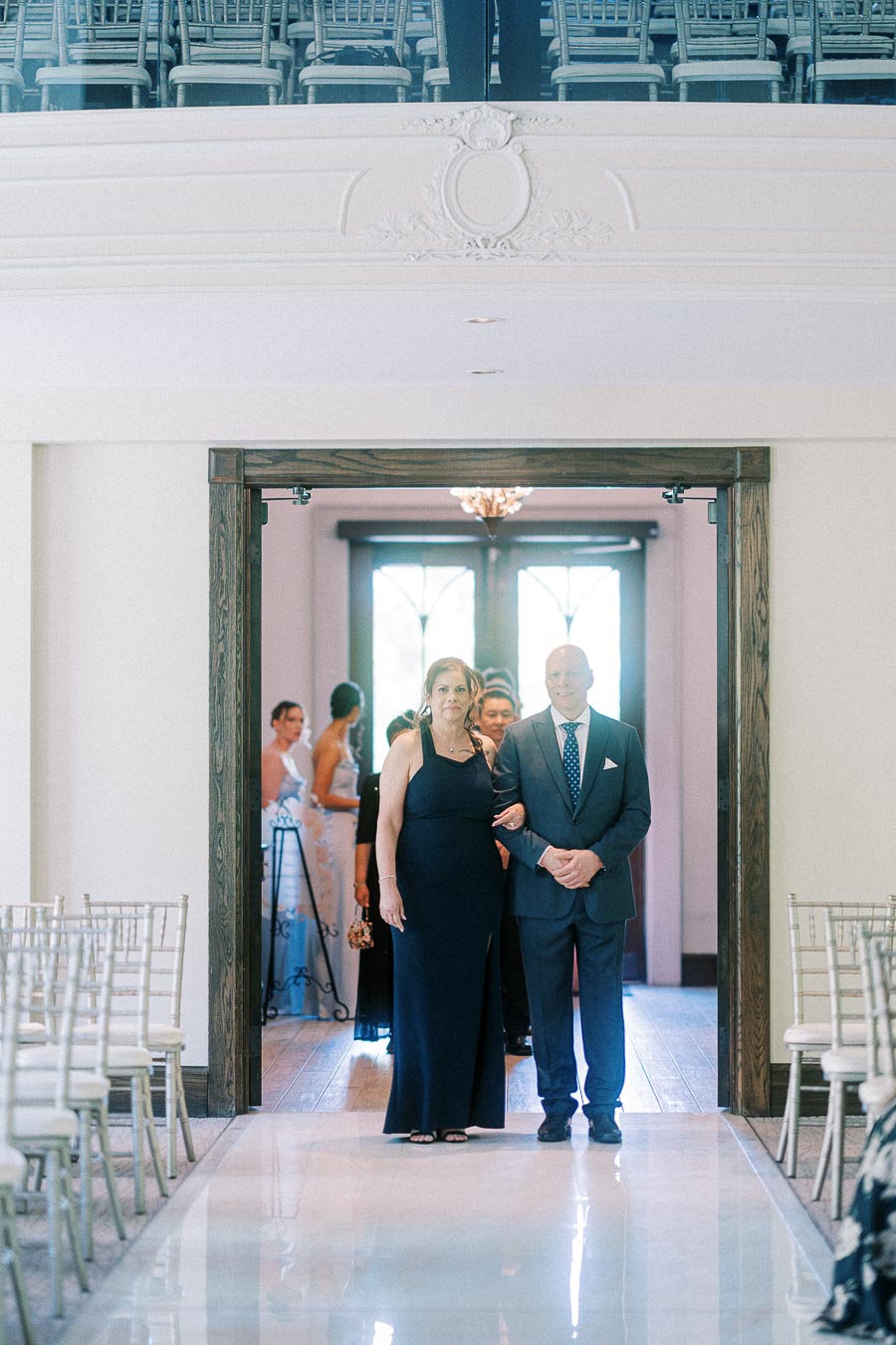 Two wedding guests walk arm in arm down a glossy chapel aisle lined with white chairs.