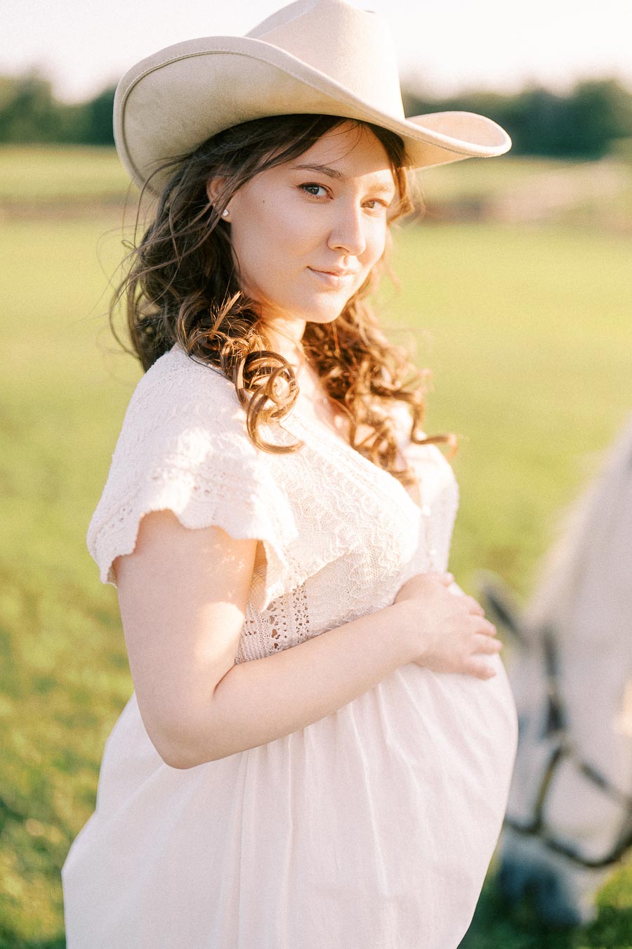 Pregnant woman in white dress and cowboy hat standing beside a horse