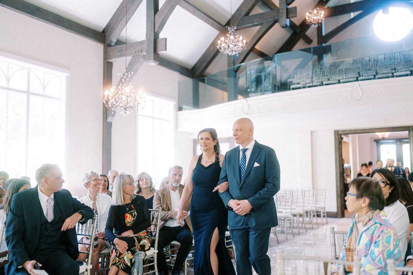A woman in a navy dress escorts a suited man down the bright chapel aisle as wedding guests watch.