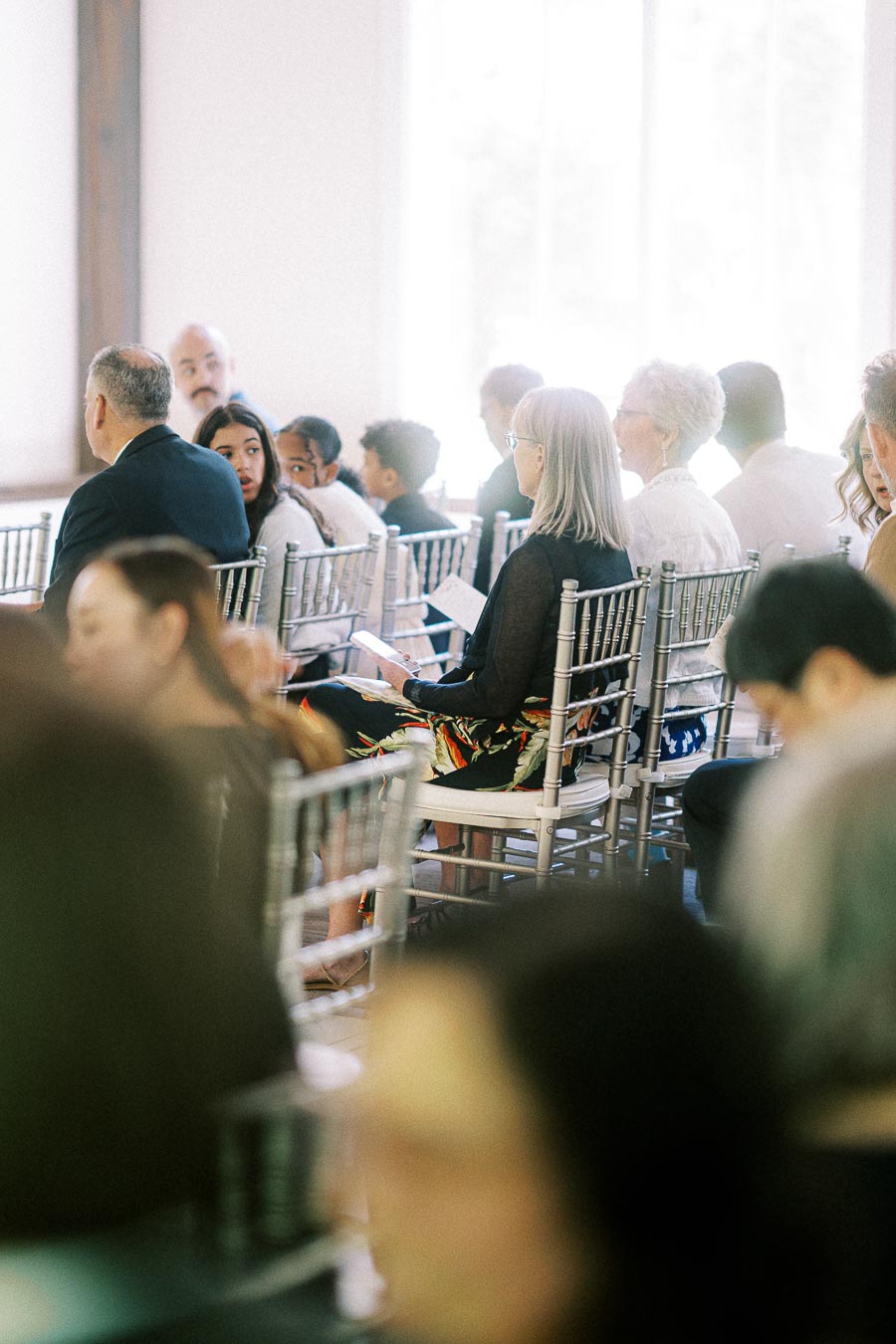 Wedding guests seated on silver chairs in a bright ceremony room, holding programs and waiting for the service.