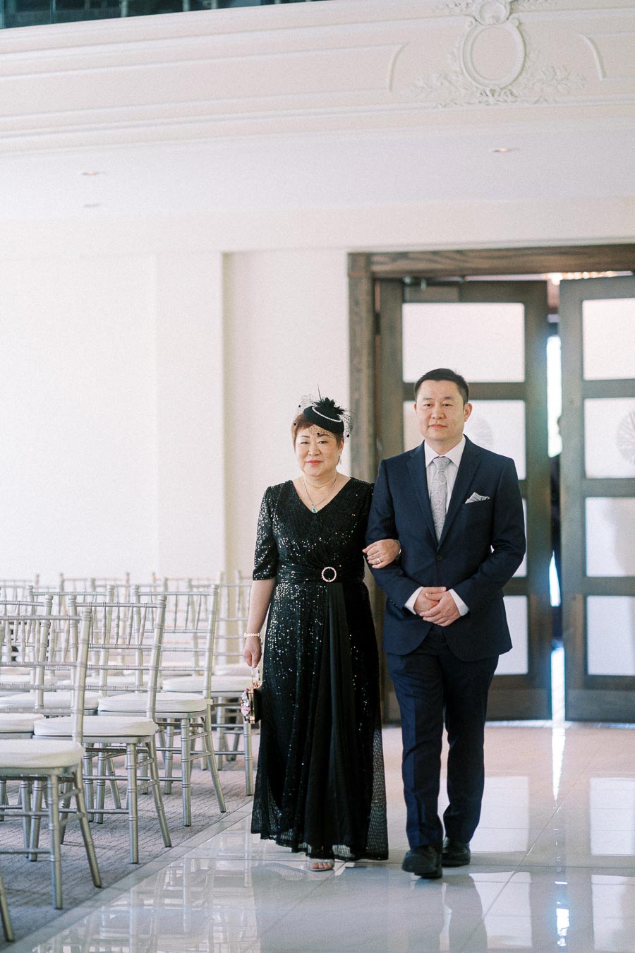 A man in a navy suit escorts a woman in a black dress down a bright wedding ceremony aisle.