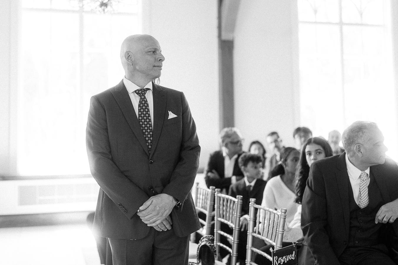Black-and-white ceremony scene with a suited man standing beside seated wedding guests in a bright room.