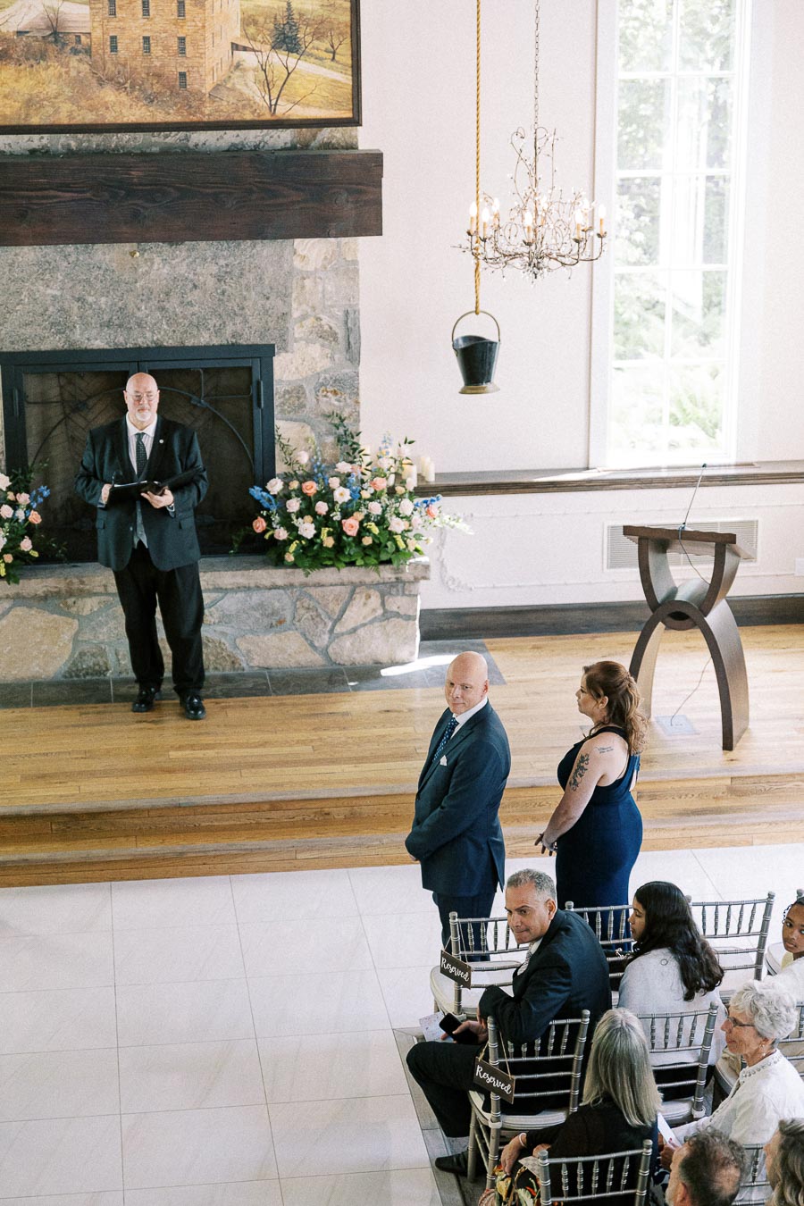 Guests stand and sit facing an officiant by a stone fireplace during an indoor wedding ceremony.