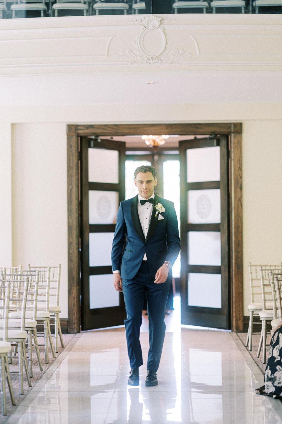Groom in a navy tuxedo walking down a bright ceremony aisle between rows of white chairs.