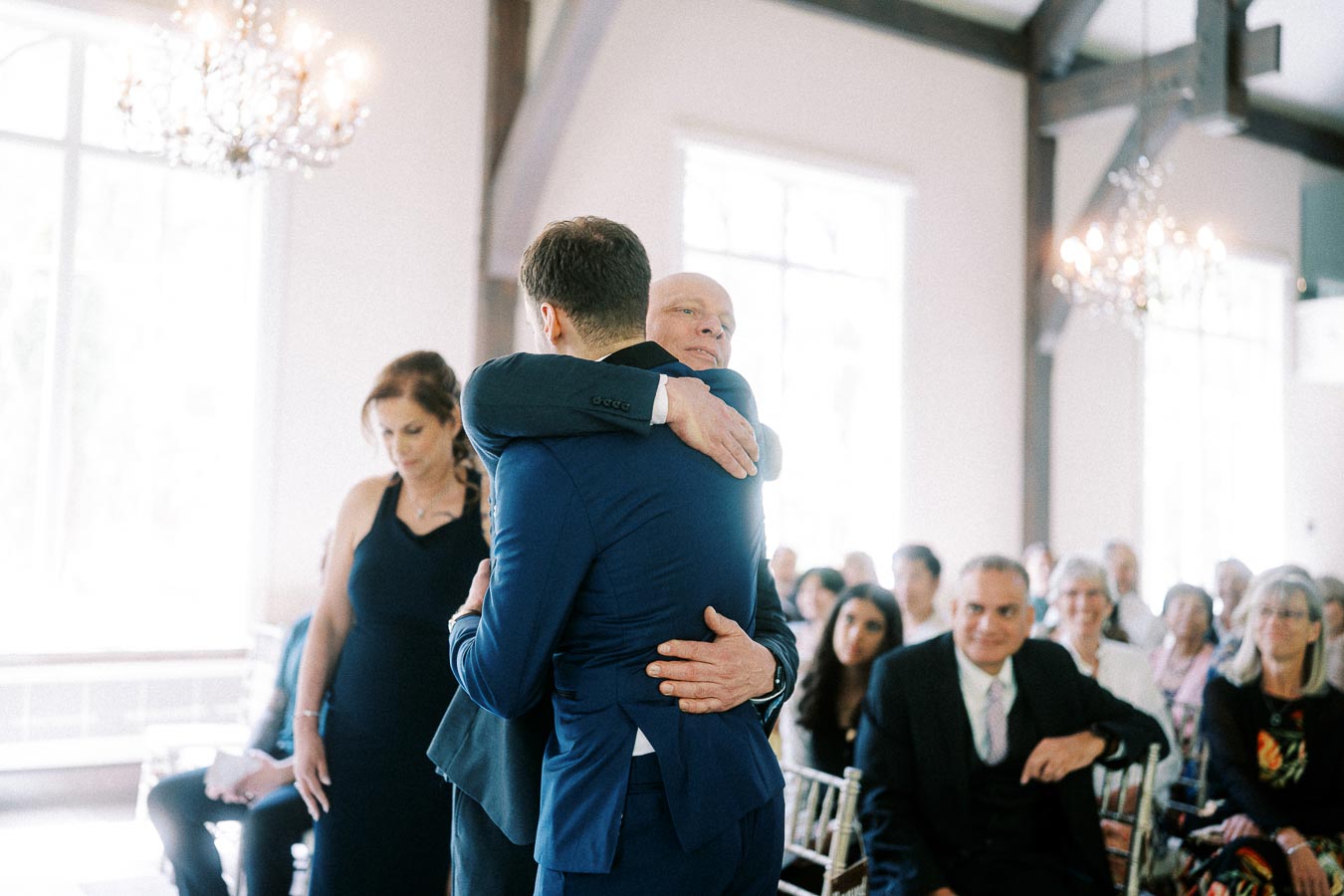 Two men in blue suits embrace during a bright indoor wedding ceremony as seated guests look on.