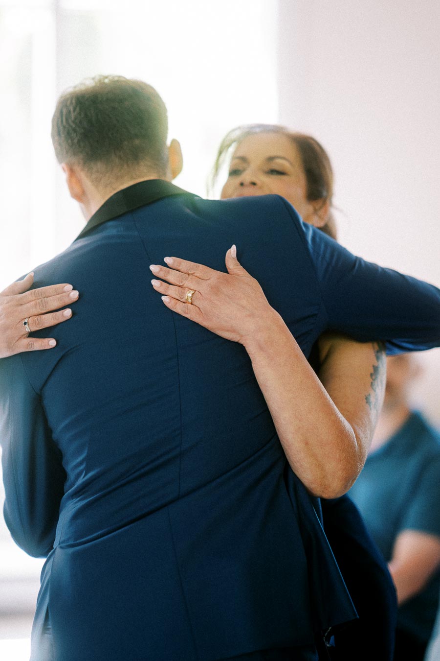 Two wedding guests embrace indoors, with hands and rings visible against a navy suit jacket.