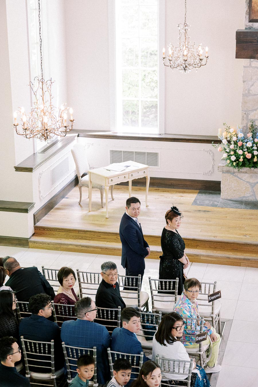 Guests seated in a bright chapel with chandeliers before Miffy and Rawle’s wedding ceremony.