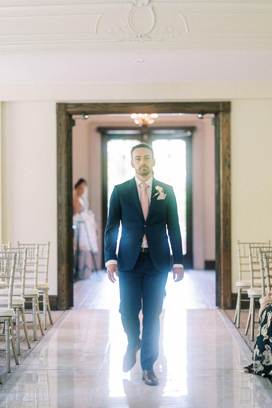 Groom in a navy suit walks down the aisle in a bright indoor ceremony room with rows of chairs.