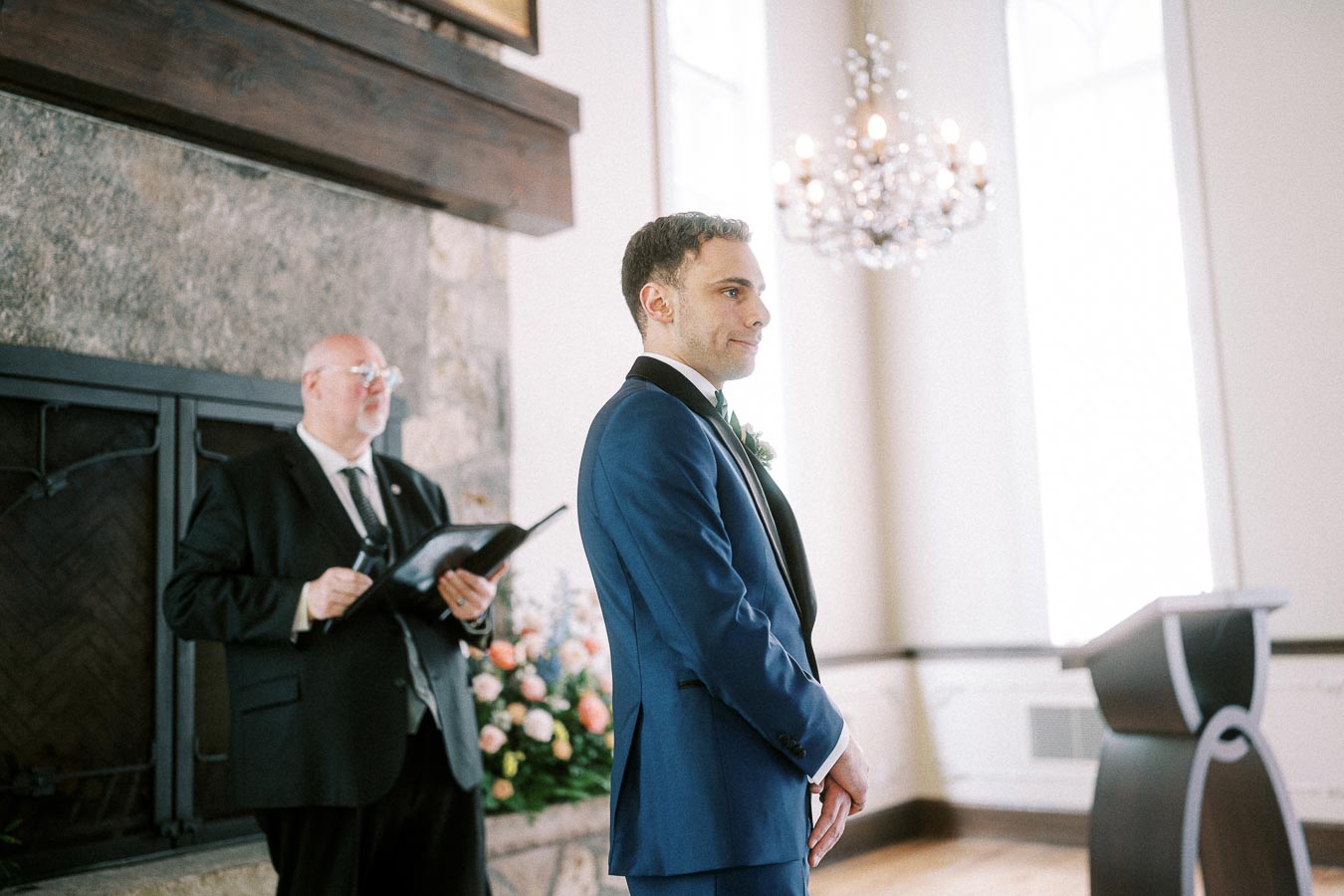 Groom in a blue tuxedo stands beside the officiant during an indoor wedding ceremony by a fireplace.