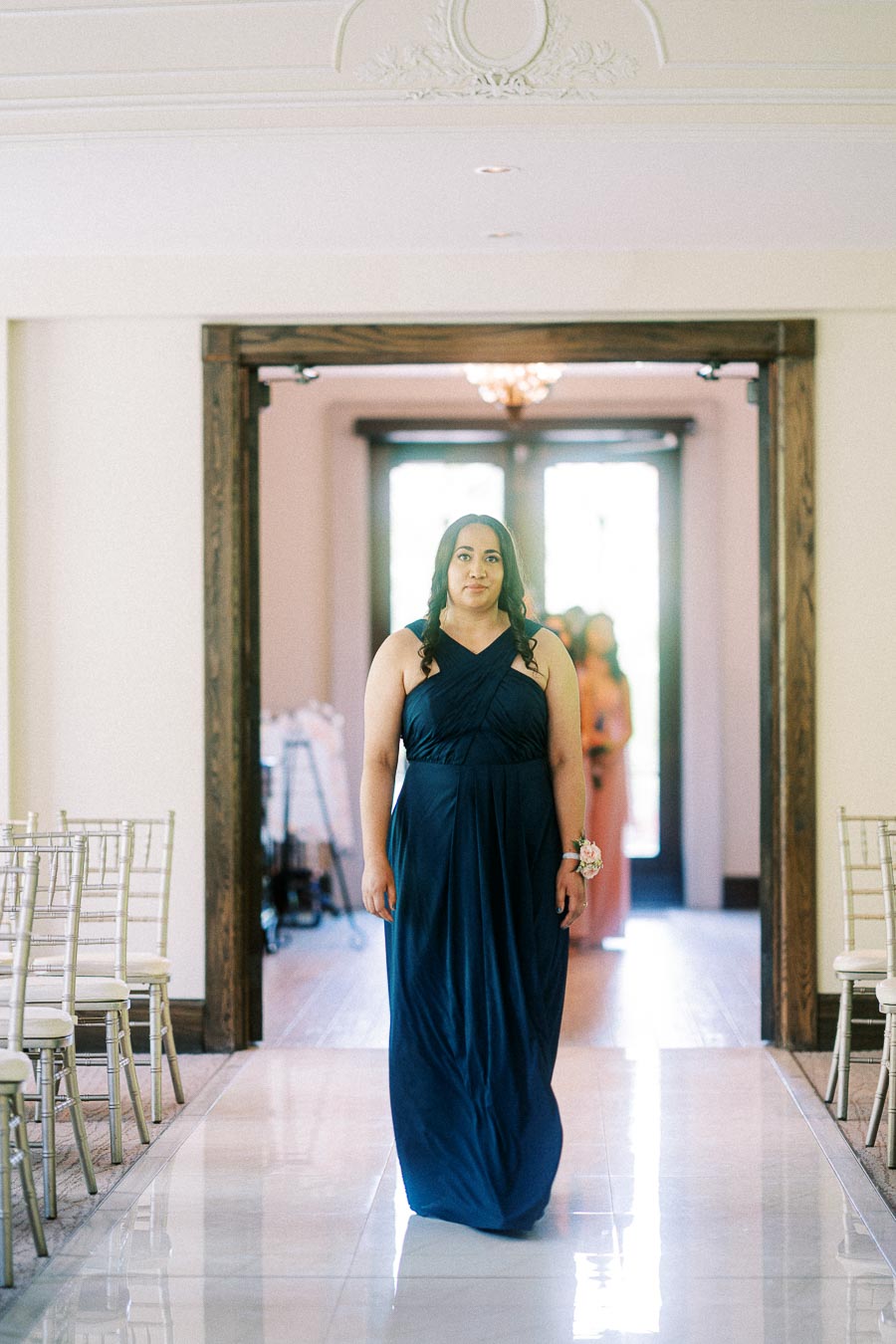Bridesmaid in a navy dress walks down a white aisle between chairs inside a wedding ceremony room.