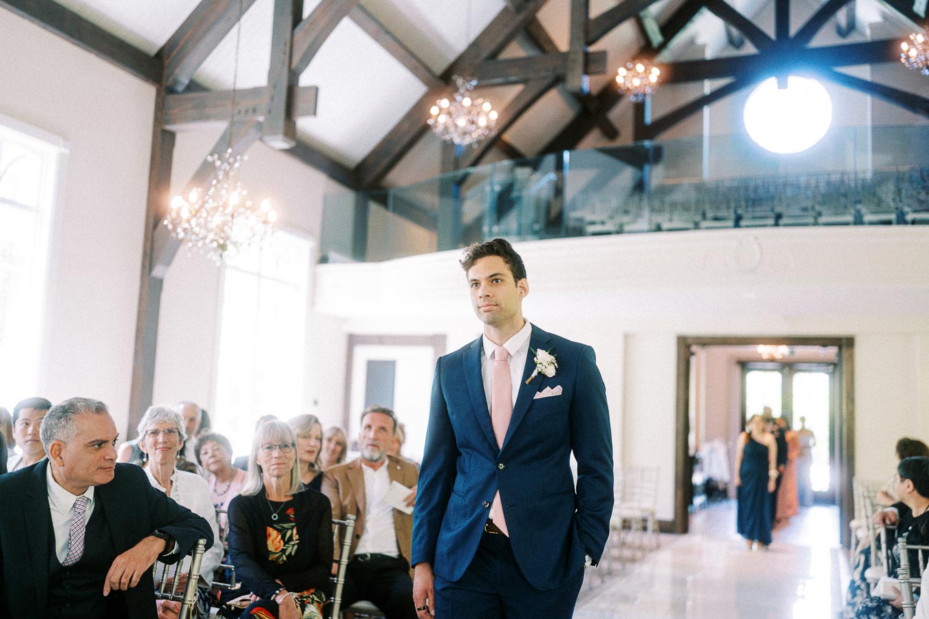 Groom in a navy suit walks down the chapel aisle as seated guests watch beneath chandeliers and timber beams.