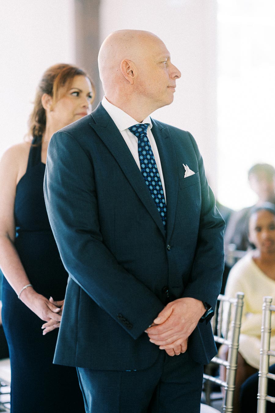 Bald man in a dark suit stands with hands clasped during an indoor wedding ceremony.