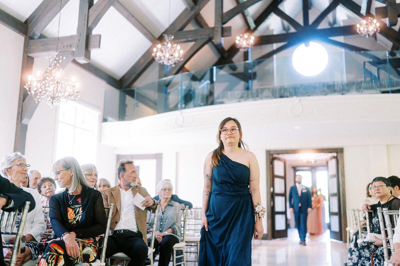 Bridesmaid in a navy dress walks down the aisle past seated guests in a bright chapel with chandeliers.