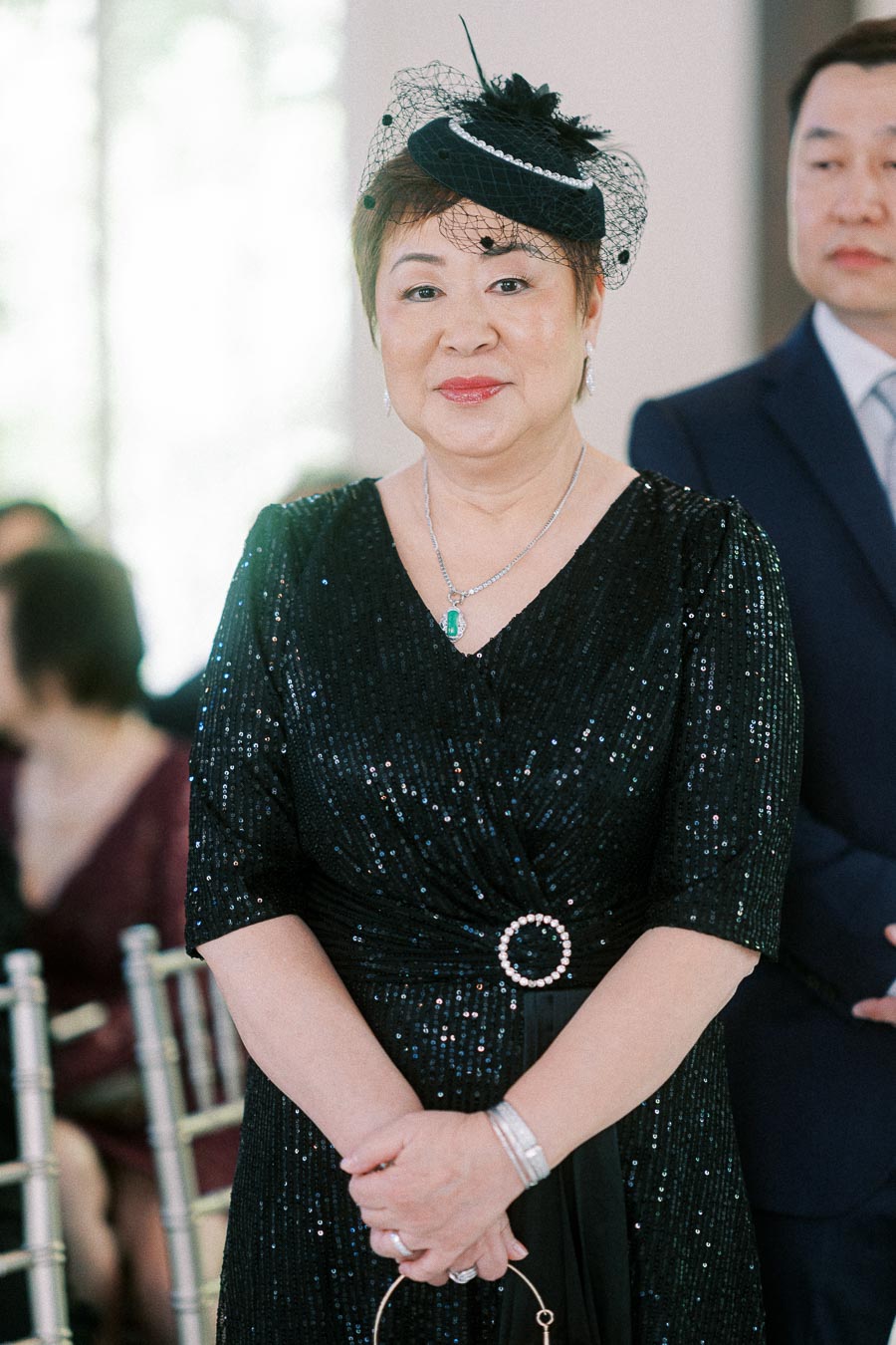 Woman in a black sequined dress and fascinator stands among seated guests during an indoor wedding ceremony.