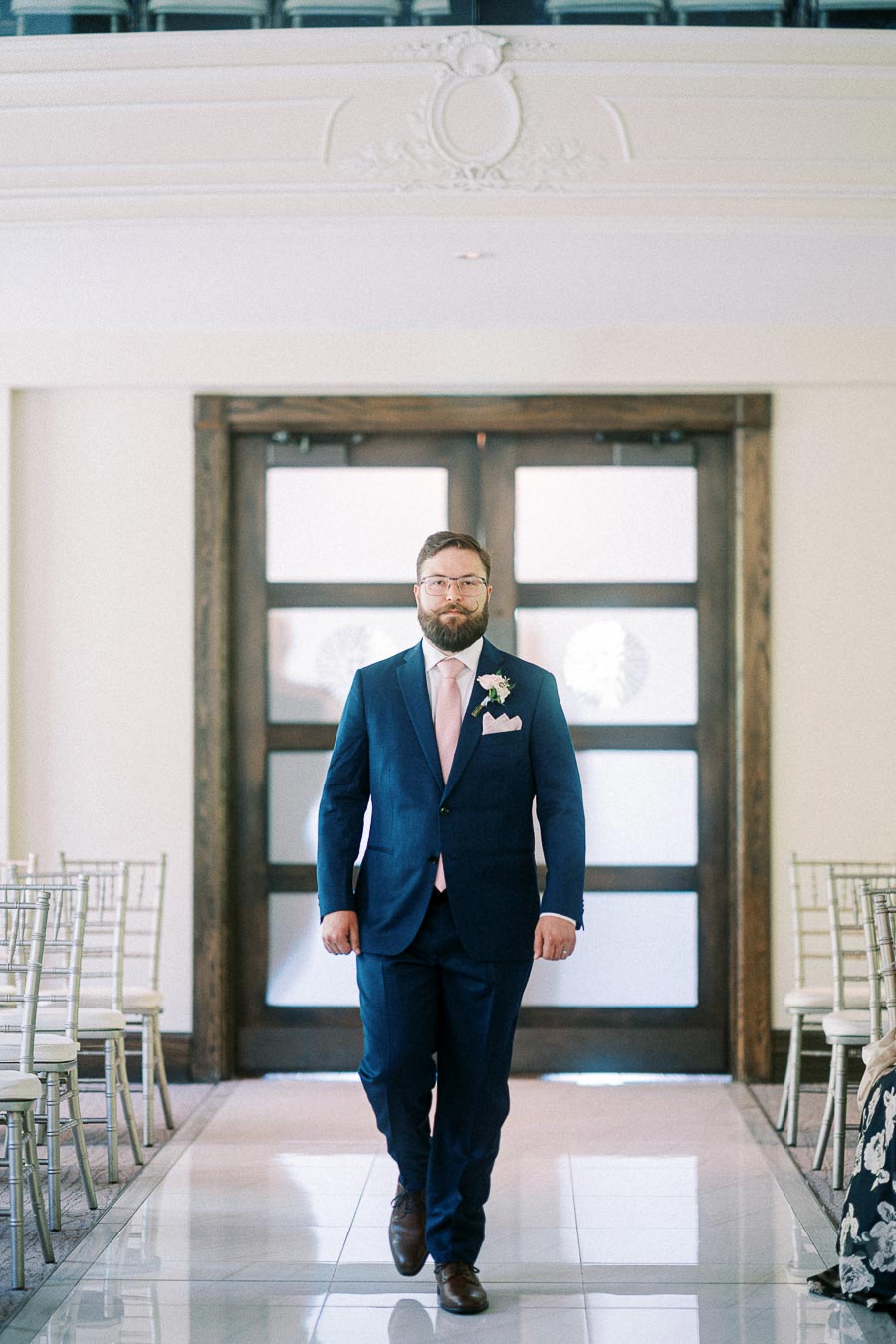Groom in a navy suit and pink tie walks down a bright chapel aisle between silver chairs.