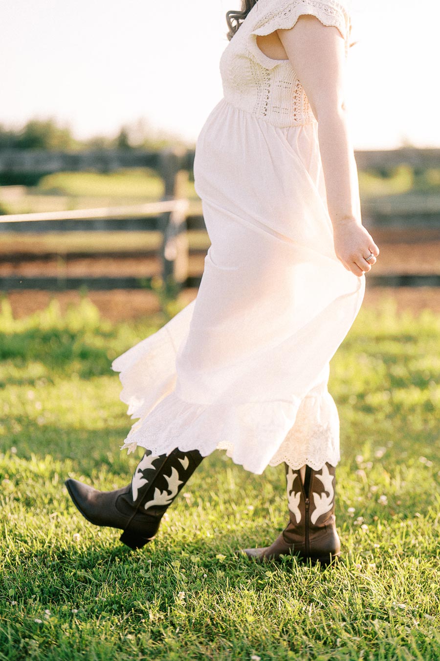 Pregnant woman in white dress and cowboy boots walking through grass