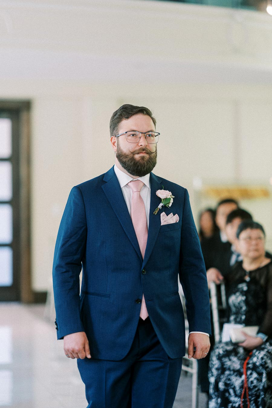Groom in a blue suit with pink tie and boutonniere walks down the ceremony aisle past seated guests.