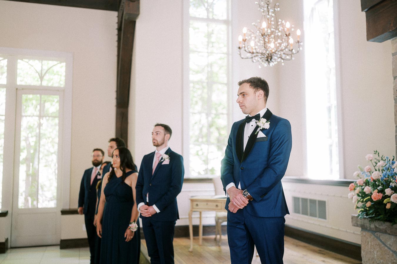 Rawle stands with attendants in a bright chapel under a chandelier during the wedding ceremony.
