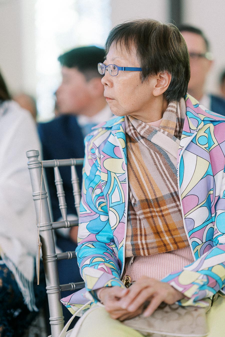 Wedding guest in blue glasses and a colorful jacket seated on a silver chair, looking to the side indoors.