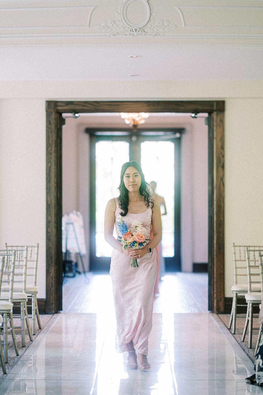 Bridesmaid in a pale pink dress walks down an indoor wedding aisle holding a colorful bouquet.
