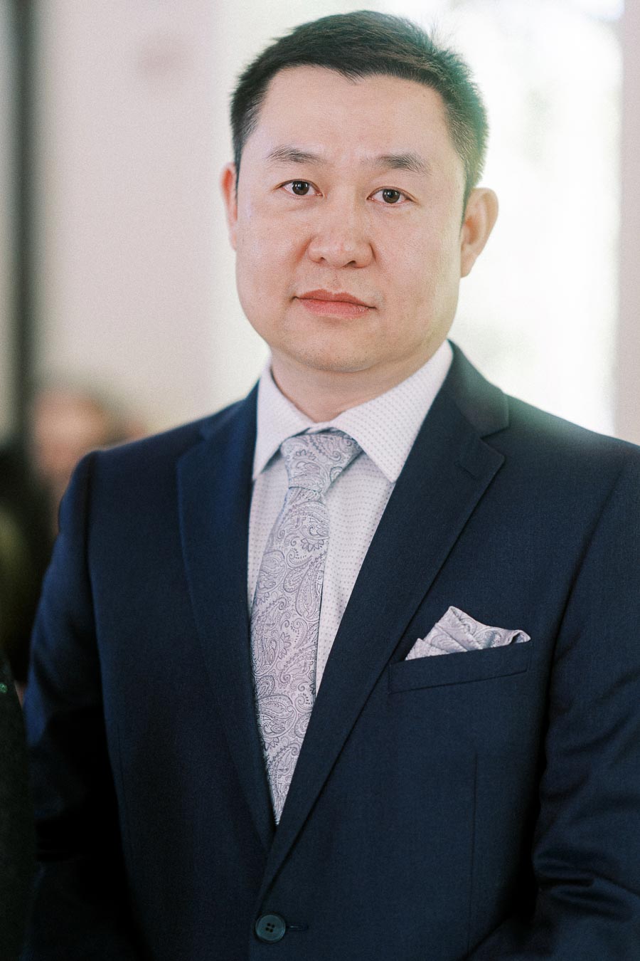 Man in a navy suit with a pale paisley tie and pocket square looking at the camera indoors.