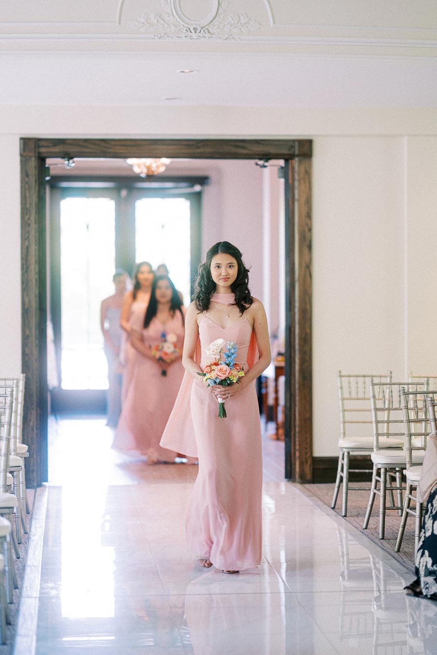 Bridesmaid in a pink dress carries a bouquet down the aisle with other bridesmaids following behind.