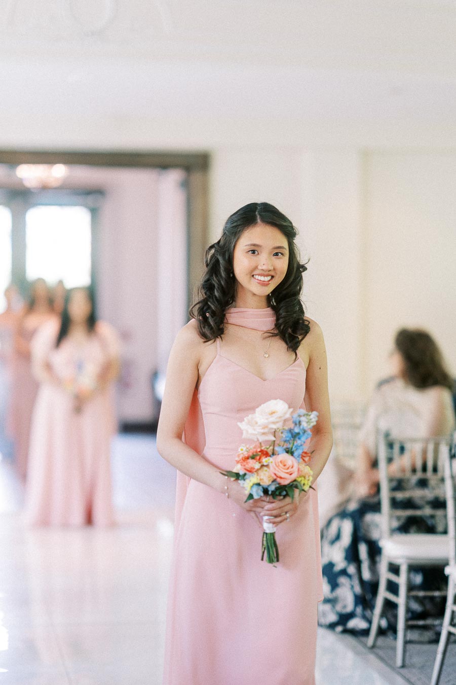 Bridesmaid in a blush pink dress holds a small bouquet while standing indoors before the ceremony.