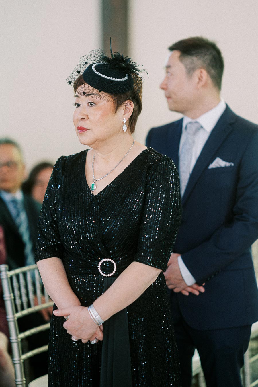 Woman in a black sequined dress and fascinator stands beside a man in a navy suit during an indoor wedding ceremony.