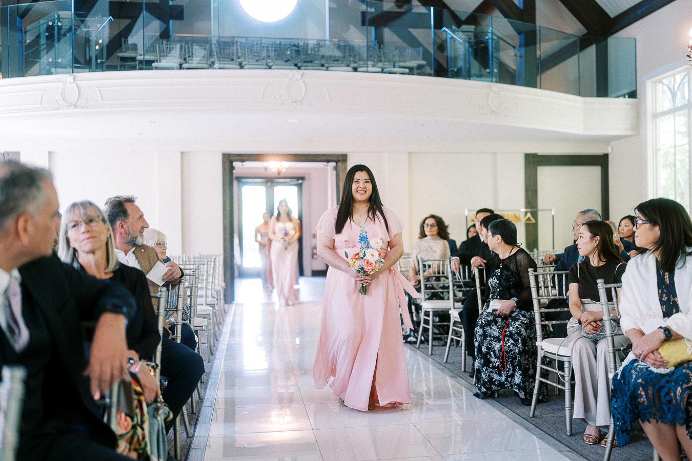 Bridesmaid in a pink dress walks down the aisle with a bouquet as seated guests watch inside Ancaster Mill.