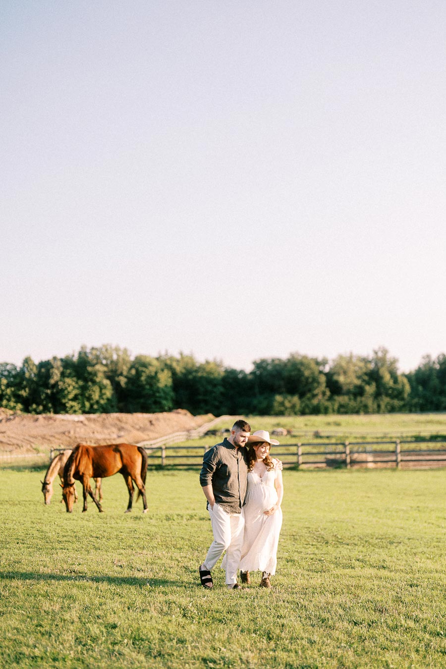 Expectant couple walking in a pasture with grazing horses at The Green Acres