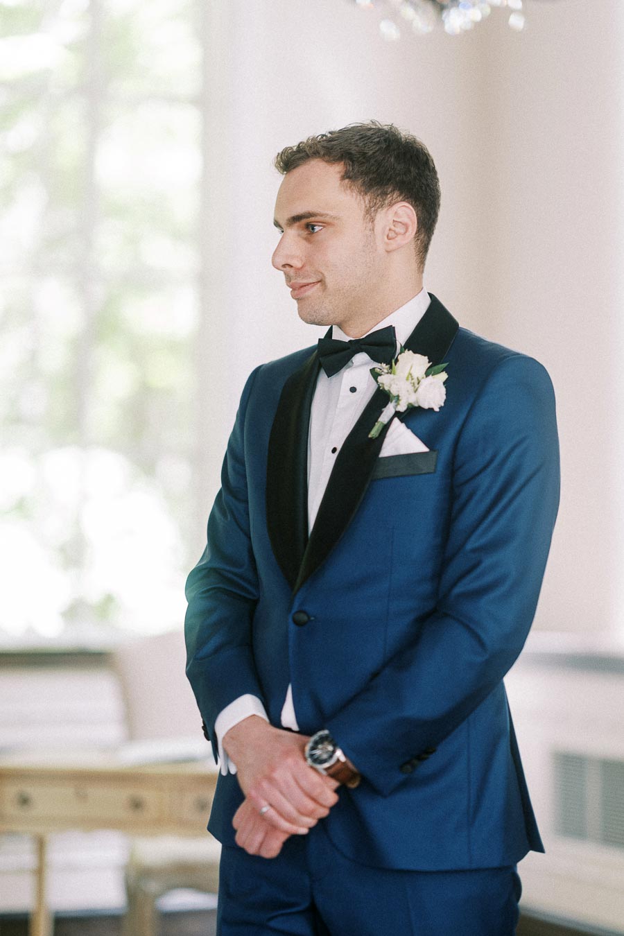 Groom in a blue tuxedo with black bow tie and white boutonniere standing indoors by a bright window.