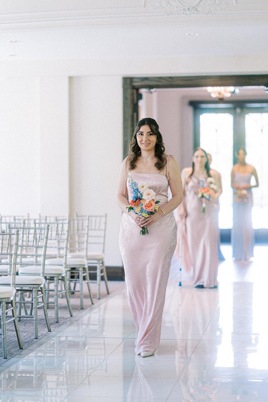 Bridesmaid in a pale pink dress walks down the aisle holding a colorful bouquet with two attendants behind her.