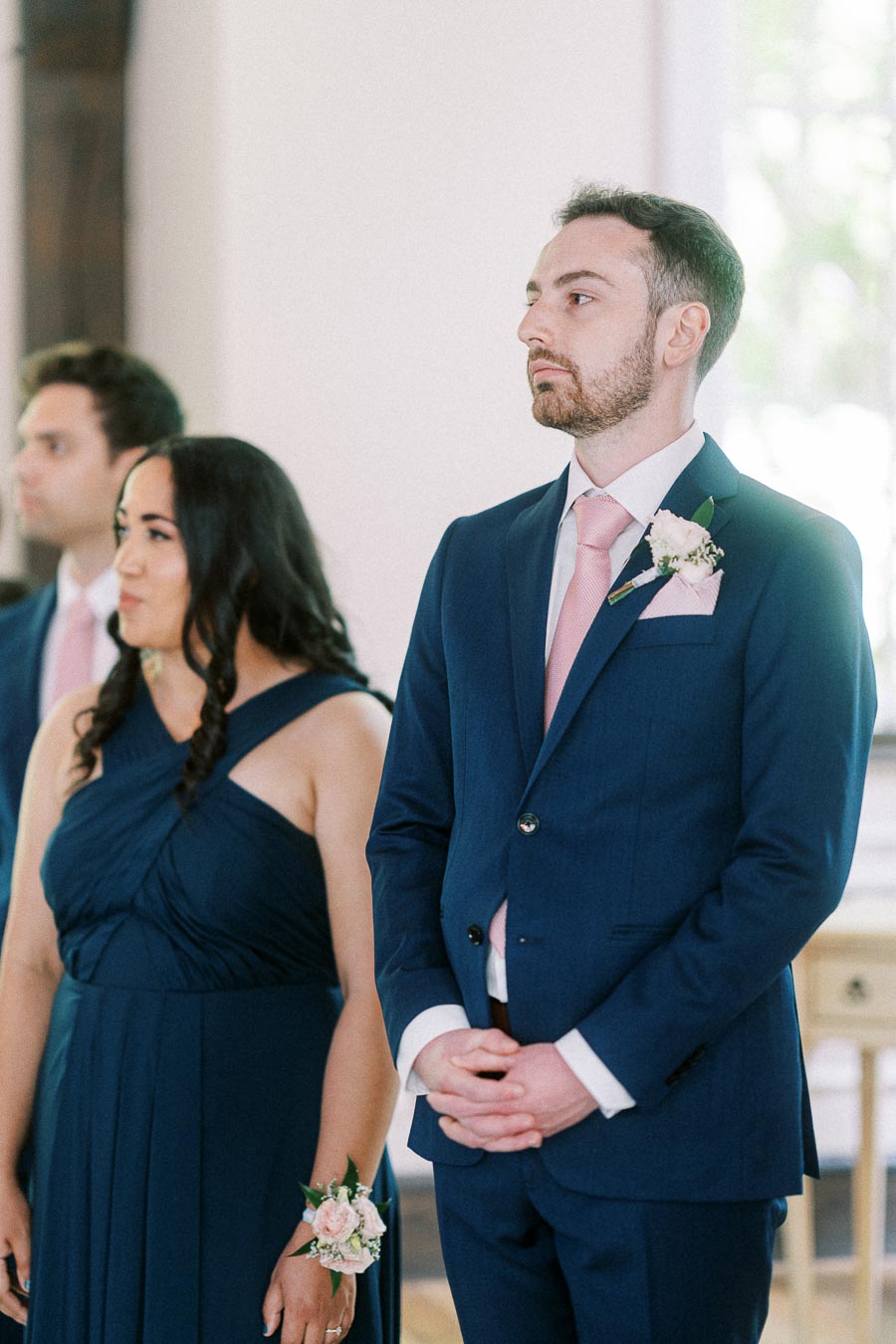 Groomsman in a navy suit with pink tie stands beside bridesmaid during an indoor wedding ceremony.