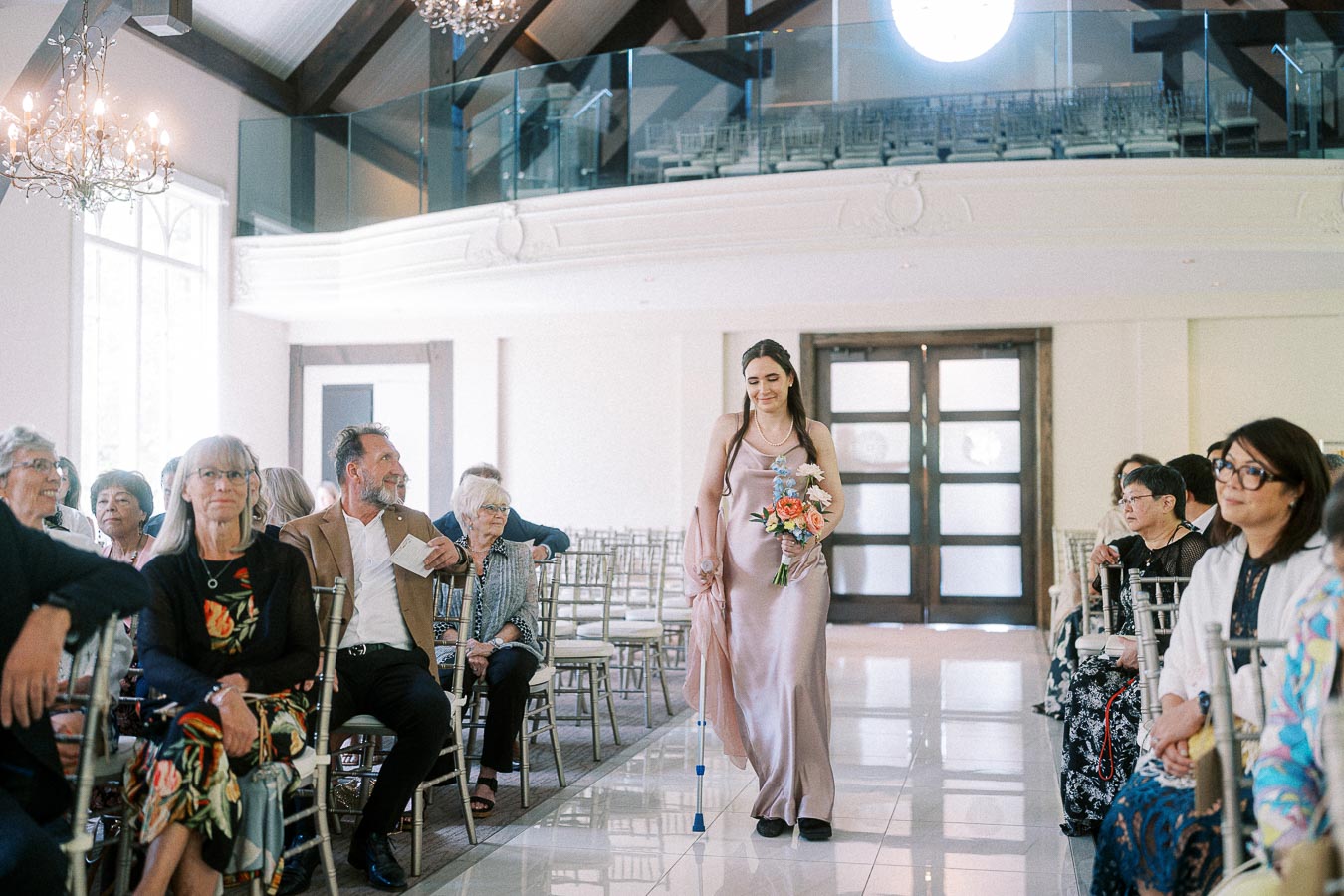 Bridesmaid in a pale pink dress walks down the aisle with a bouquet as guests watch in a bright chapel.